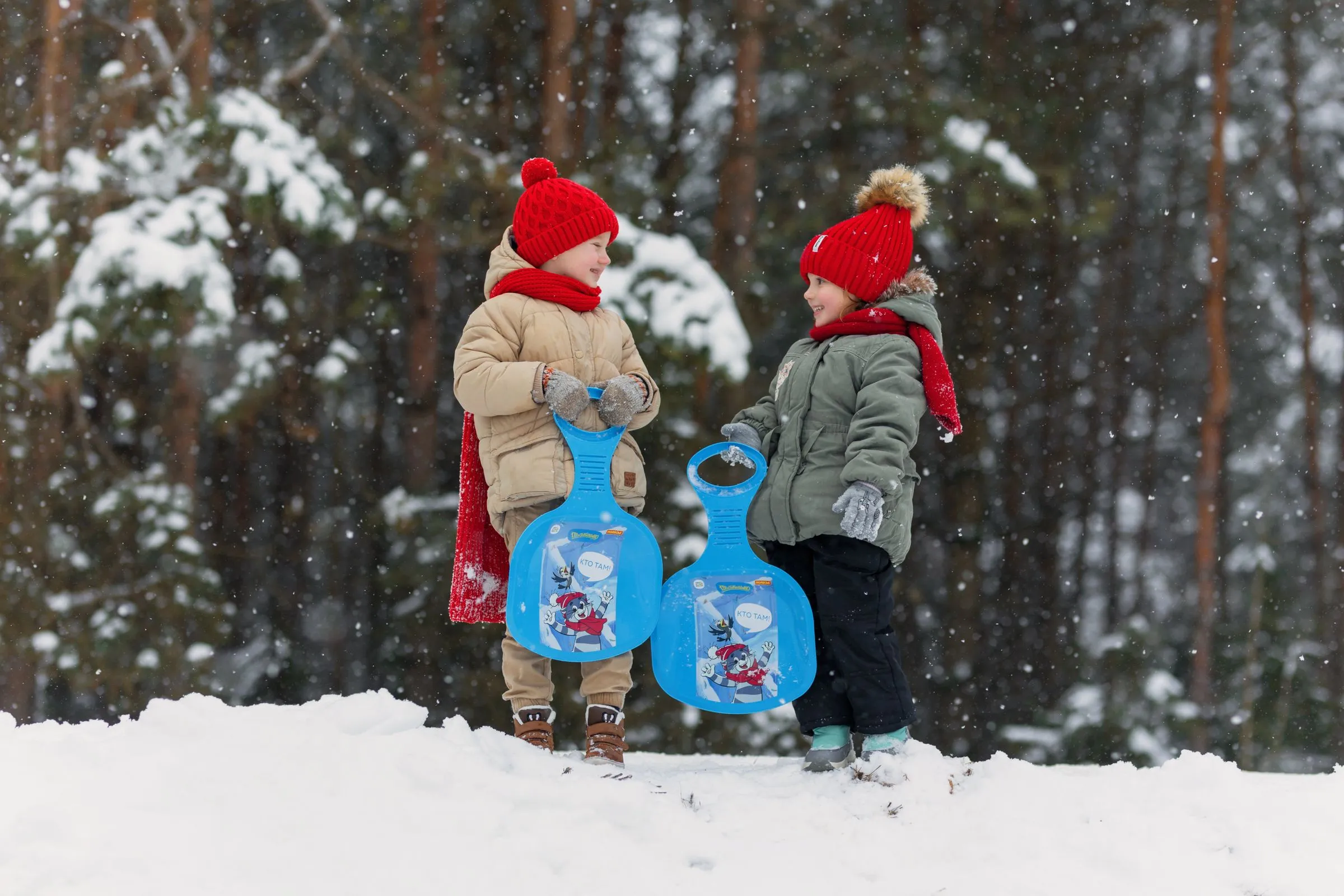Two children in red hats holding blue sleds in a snowy spruce forest